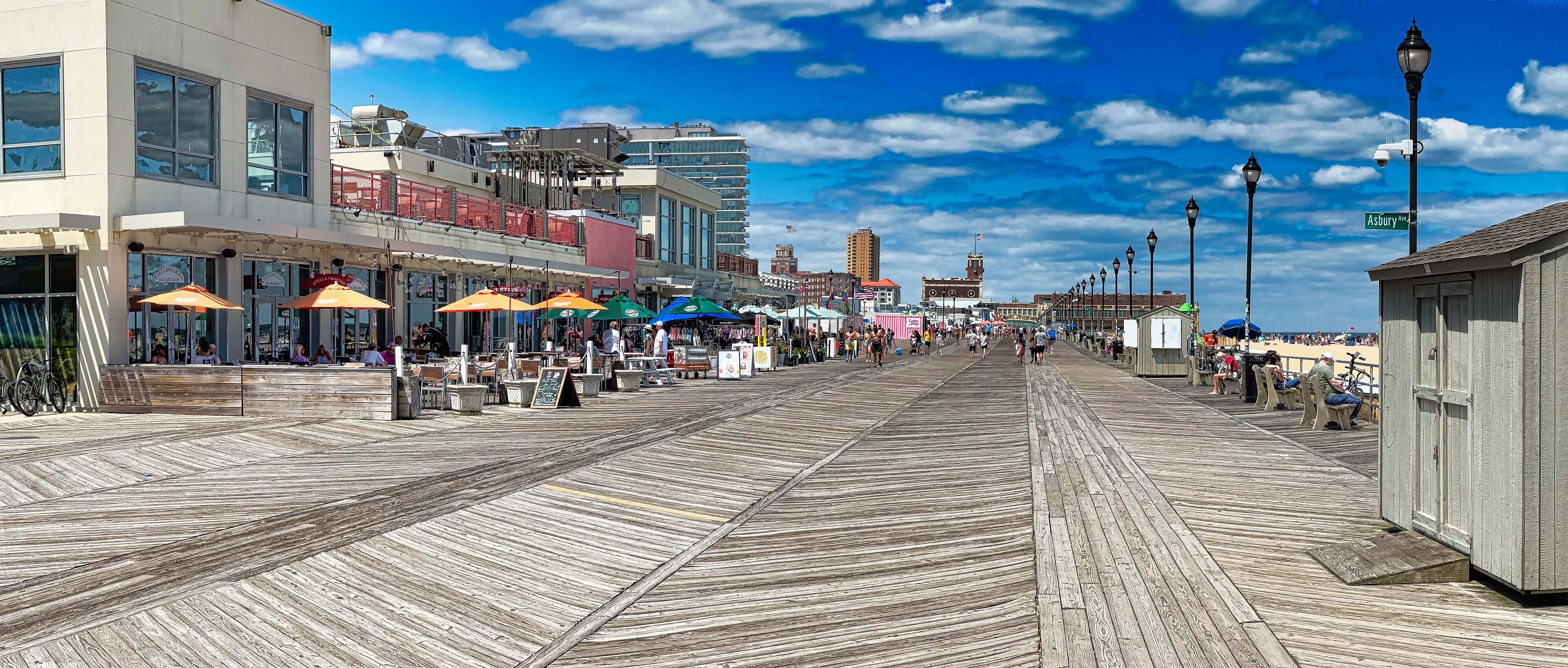 The Asbury Park boardwalk stretching ahead past umbrella-topped cafes under a blue sky with scattered clouds.