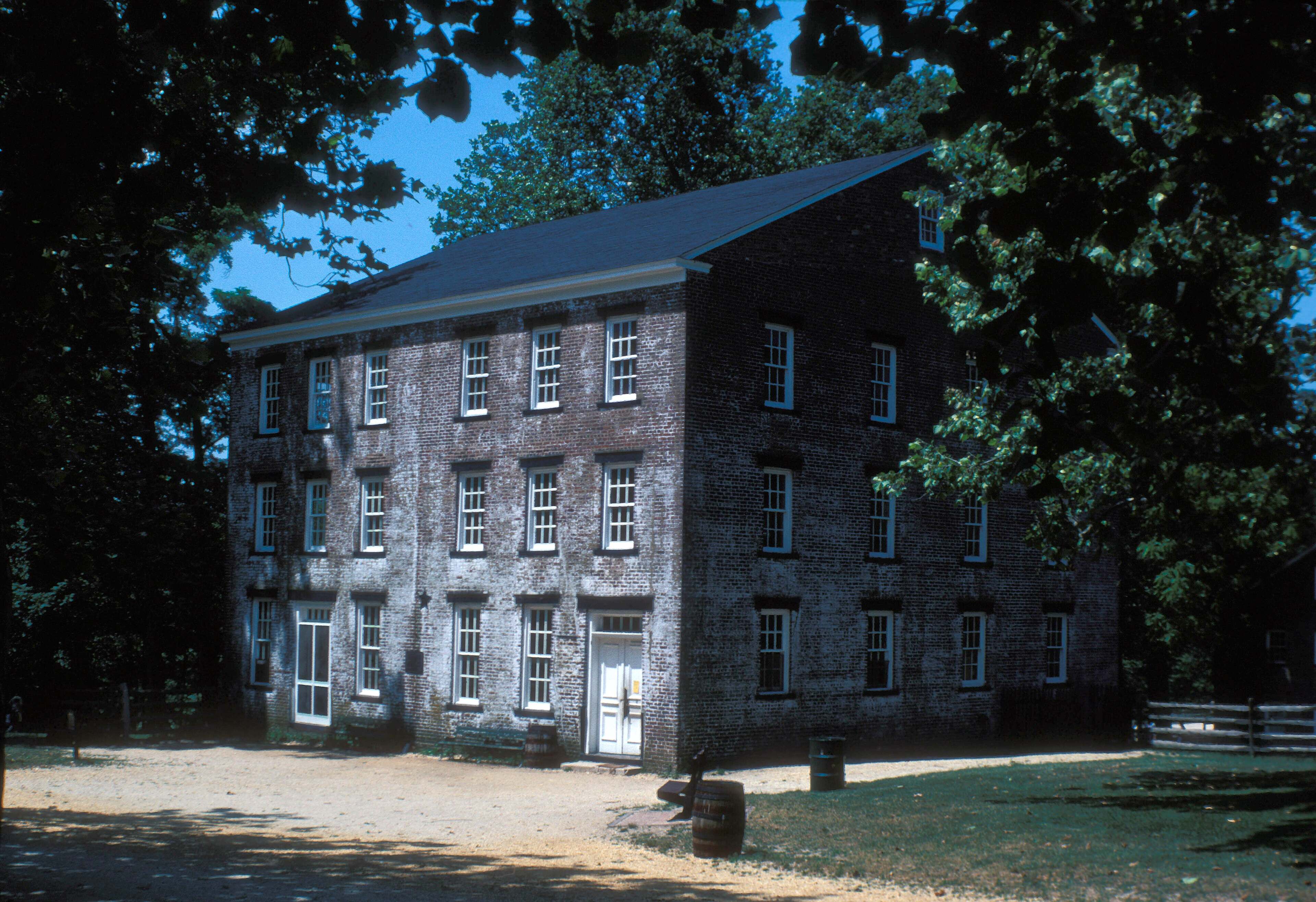 A three-story brick building at Allaire Village under tall trees, with a split-rail fence at the right.