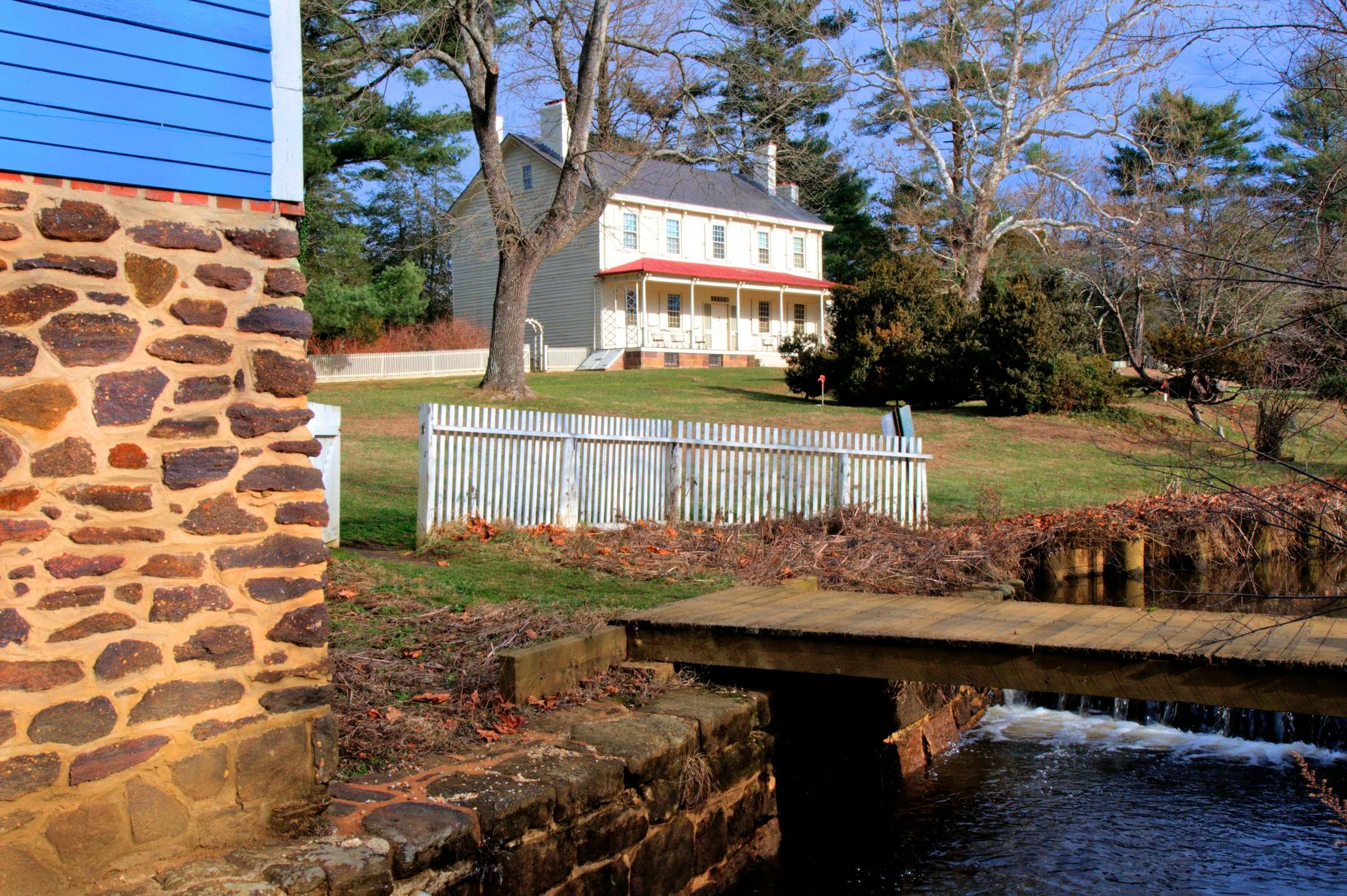 A white farmhouse beyond a mill race at Historic Walnford, with a fieldstone wall in the foreground.
