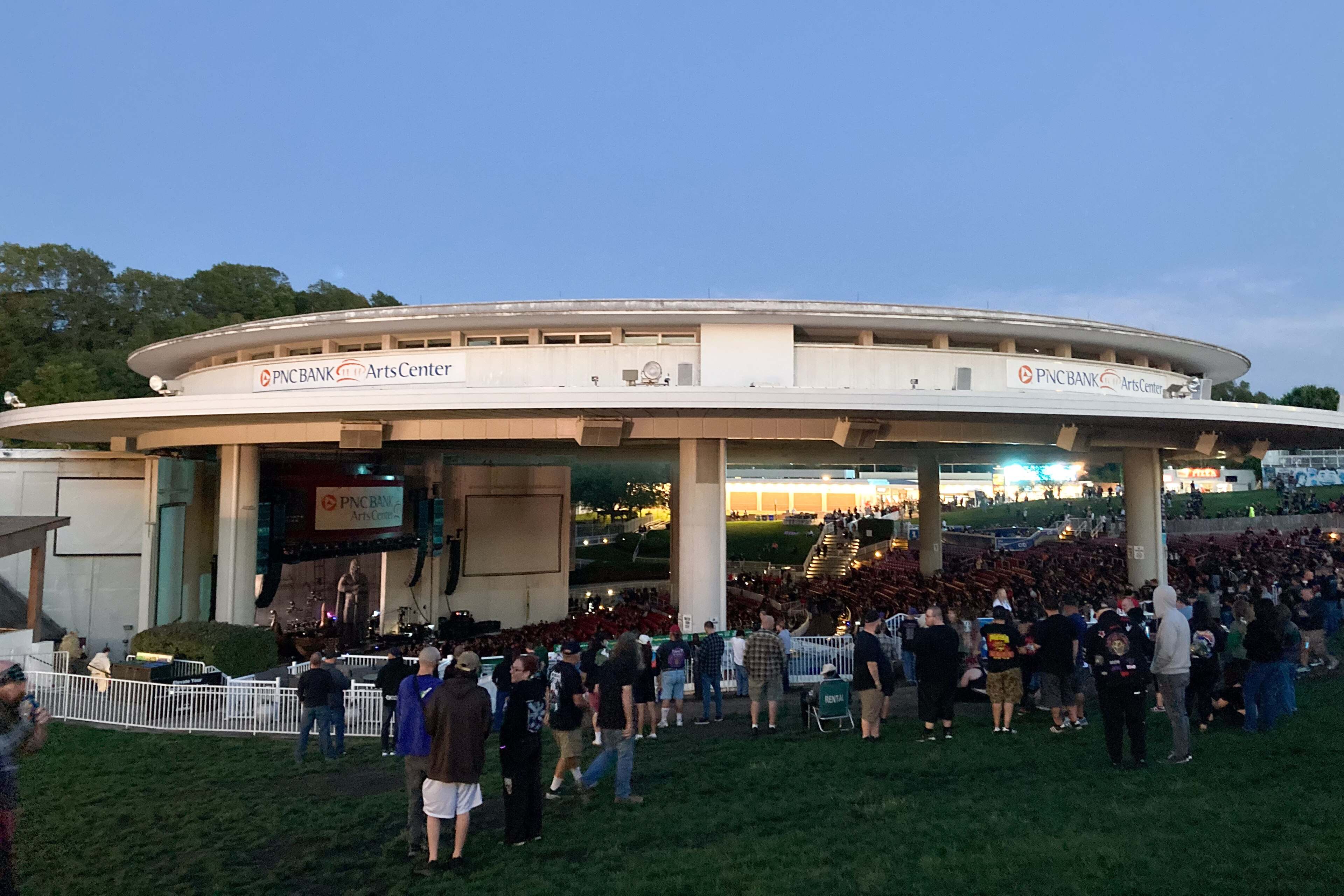 The circular roof of the PNC Bank Arts Center over the stage and seating lawn at dusk, with a crowd on the grass.