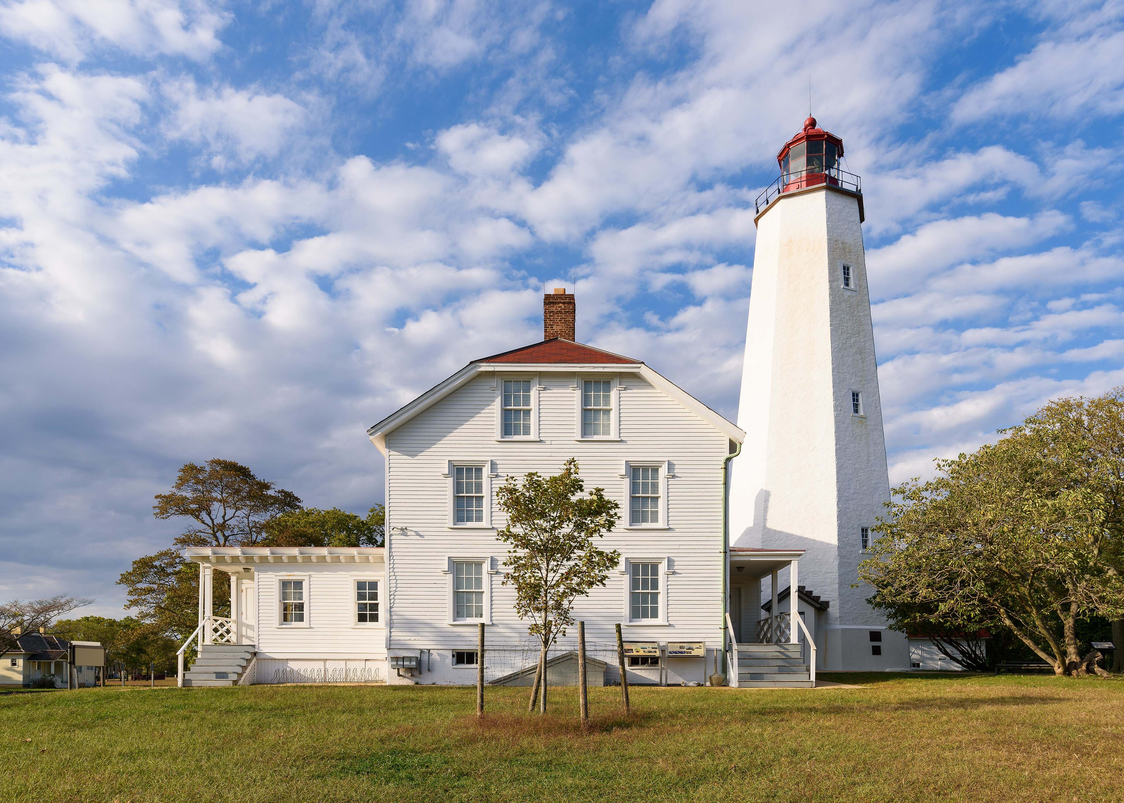 The white Sandy Hook Lighthouse tower and keeper's house under a sky of scattered clouds.