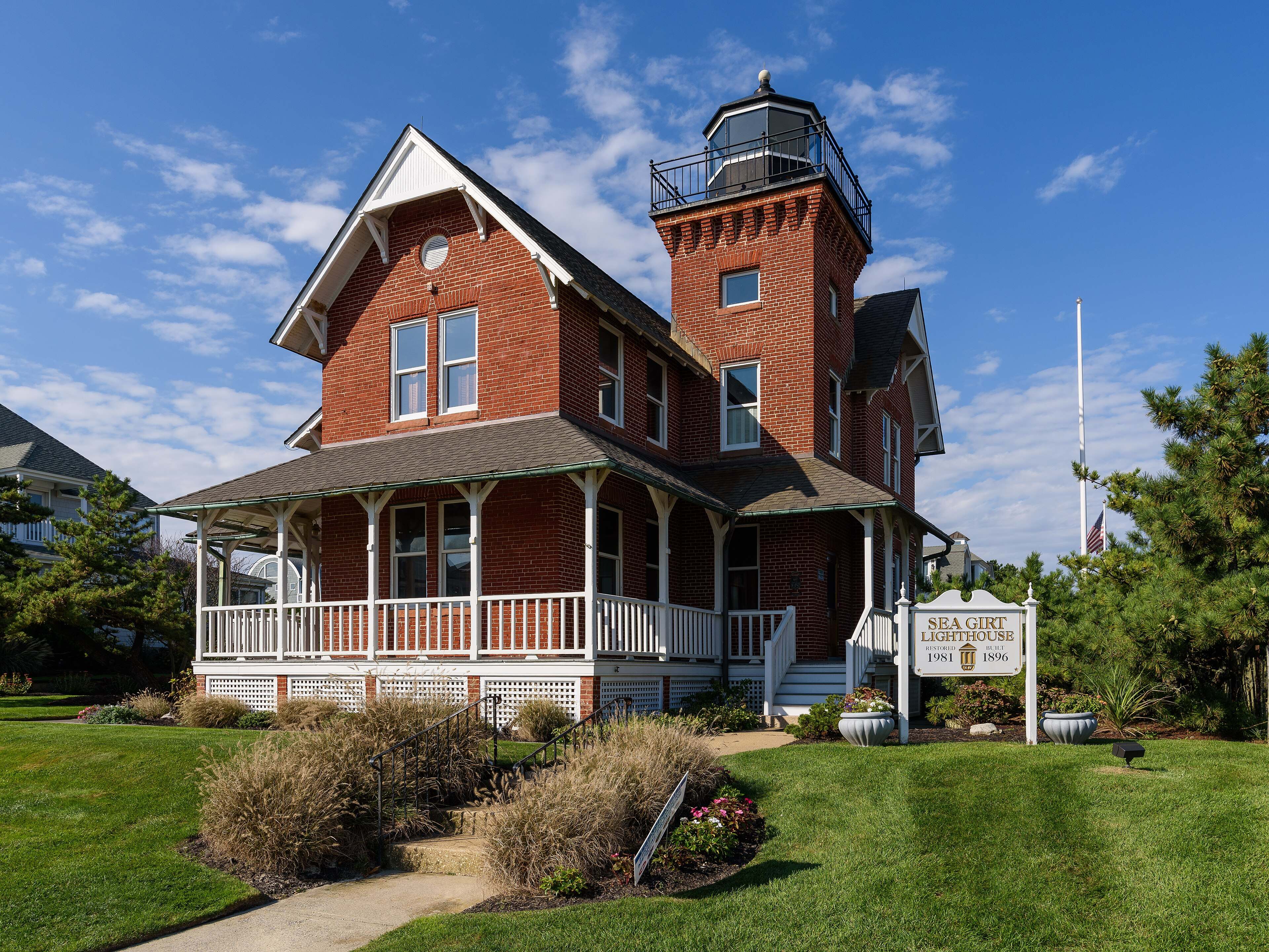 The red-brick Sea Girt Lighthouse with its wraparound porch and square lantern tower, behind a garden and lawn.
